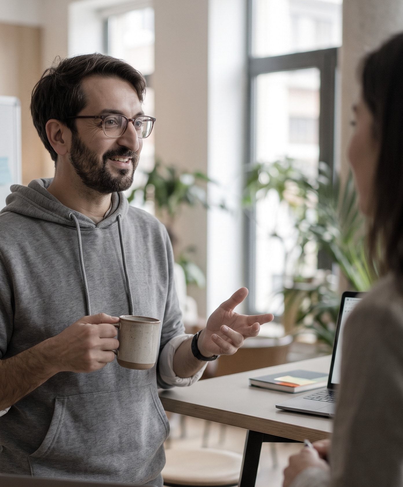 Namir in a grey hoodie, holding a coffee mug and talking with a colleague, a bright window and plants behind him