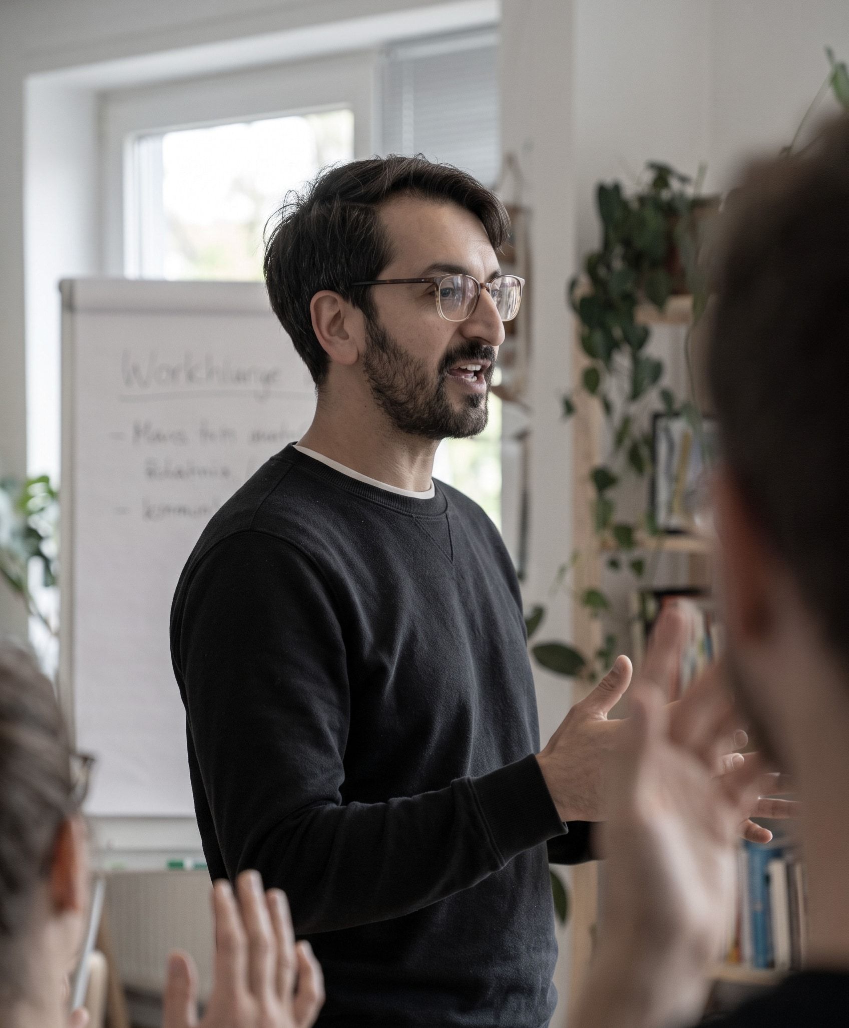 Namir im schwarzen Sweatshirt, mitten im Satz vor einer Gruppe, hinter ihm ein Flipchart und Pflanzen